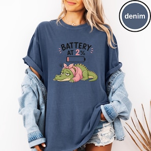 May include: A denim blue t-shirt featuring a graphic of a tired green crocodile wearing a pink headscarf. Above the crocodile is a low battery icon and the text BATTERY AT 2%. The shirt has short sleeves and a crew neckline, shown with a denim jacket.
