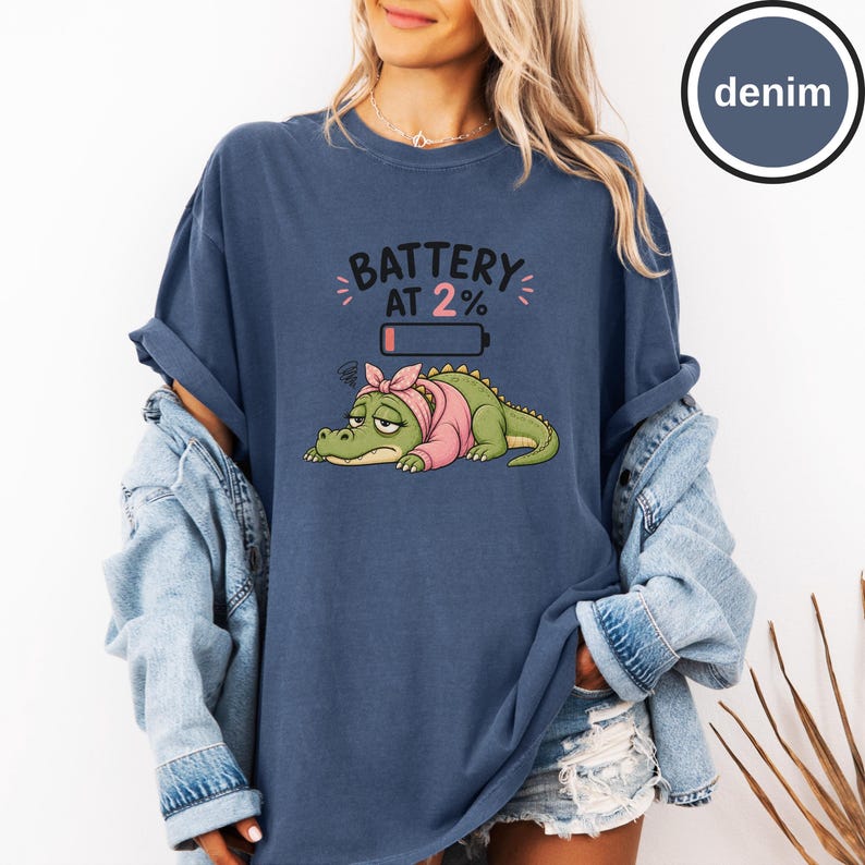 May include: A denim blue t-shirt featuring a graphic of a tired green crocodile wearing a pink headscarf. Above the crocodile is a low battery icon and the text BATTERY AT 2%. The shirt has short sleeves and a crew neckline, shown with a denim jacket.