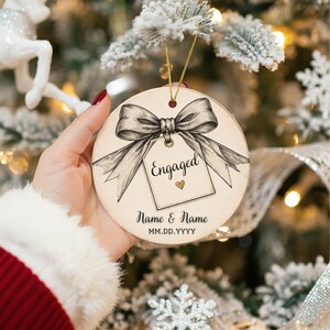 May include: A round, cream-colored ceramic ornament featuring a black-ink illustration of a large bow tied to a gift tag. The tag displays the word Engaged with a small gold heart. Below, the text reads Name & Name and MM.DD.YYYY.