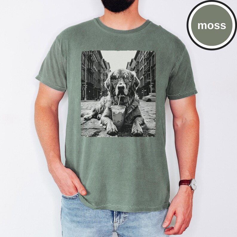 Peut inclure: Un t-shirt &agrave; manches courtes vert mousse avec une impression graphique en noir et blanc repr&eacute;sentant un chien portant des lunettes de soleil et buvant dans un gobelet en plastique avec une paille, sur fond de rue urbaine bord&eacute;e d'immeubles.