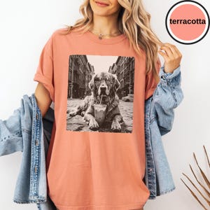 Peut inclure: Un t-shirt couleur terre cuite avec une impression graphique en noir et blanc repr&eacute;sentant un chien portant des lunettes de soleil et buvant dans un gobelet avec une paille, sur fond de rue urbaine. Le t-shirt a un col rond et des manches courtes.