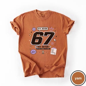 Pu&ograve; includere: Una t-shirt color ruggine con una grande grafica nera del numero 67 e il testo SIX SEVEN, NOT JUST A NUMBER. IT'S A MOOD. Include grafiche di una faccina sorridente, corona, clessidra, stella, cursore e una nota 67% CHAOS 33% CONFIDENCE.