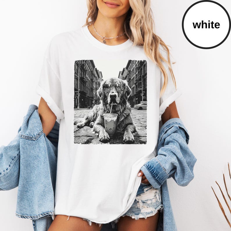 Peut inclure: Un t-shirt blanc avec une impression graphique en noir et blanc repr&eacute;sentant un chien portant des lunettes de soleil et buvant dans un gobelet avec une paille, allong&eacute; dans une rue. Le t-shirt a des manches courtes et un col rond.