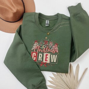 Op de afbeelding: Een olijfgroene sweater met ronde hals en een grafische print van versierde kerstbomen en de tekst "Christmas CREW" in rode en witte blokletters. Het ontwerp bevat rode strikken en steraccenten, met een bruine hoed in de buurt.