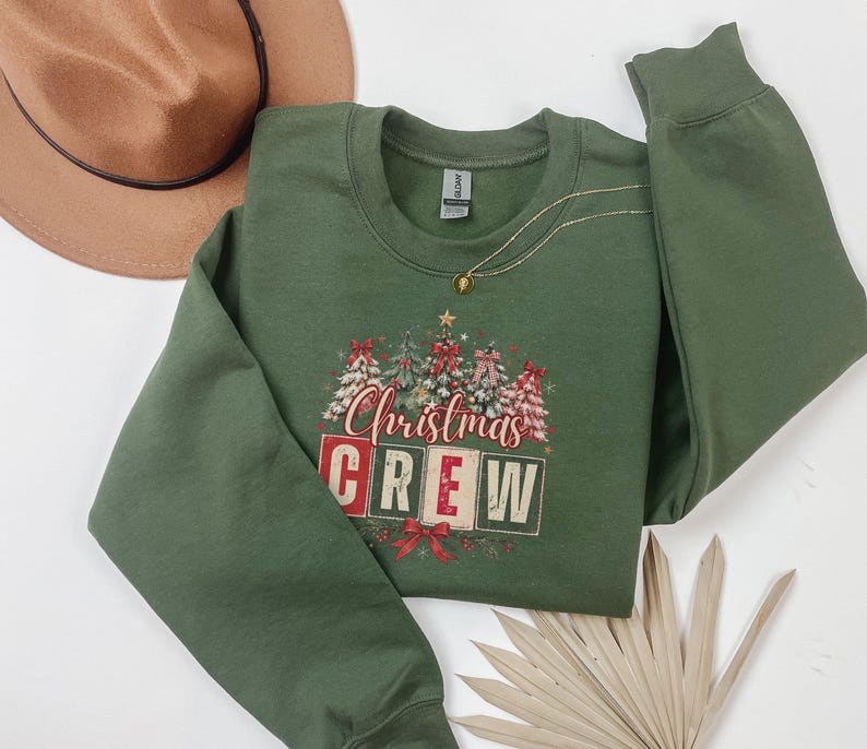 Op de afbeelding: Een olijfgroene sweater met ronde hals en een grafische print van versierde kerstbomen en de tekst "Christmas CREW" in rode en witte blokletters. Het ontwerp bevat rode strikken en steraccenten, met een bruine hoed in de buurt.