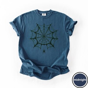 May include: A blue short-sleeve t-shirt featuring a dark green graphic of a spider web centered on the chest. A small spider hangs from the bottom of the web. The fabric has a slightly washed, textured appearance with a crew neckline.