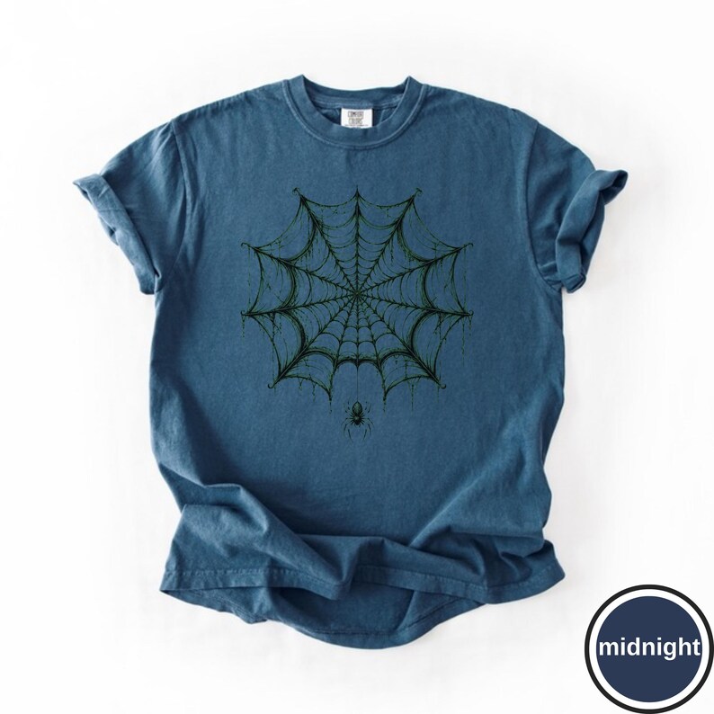 May include: A blue short-sleeve t-shirt featuring a dark green graphic of a spider web centered on the chest. A small spider hangs from the bottom of the web. The fabric has a slightly washed, textured appearance with a crew neckline.