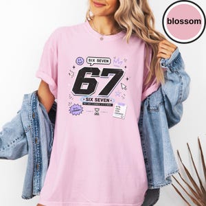 Pu&ograve; includere: Una t-shirt a maniche corte rosa chiaro con una grande grafica nera del numero 67. Il design include il testo SIX SEVEN, NOT JUST A NUMBER. IT'S A MOOD, 6+7= LEGENDARY e 67% CHAOS 33% CONFIDENCE, insieme a piccole icone come una faccina sorridente.
