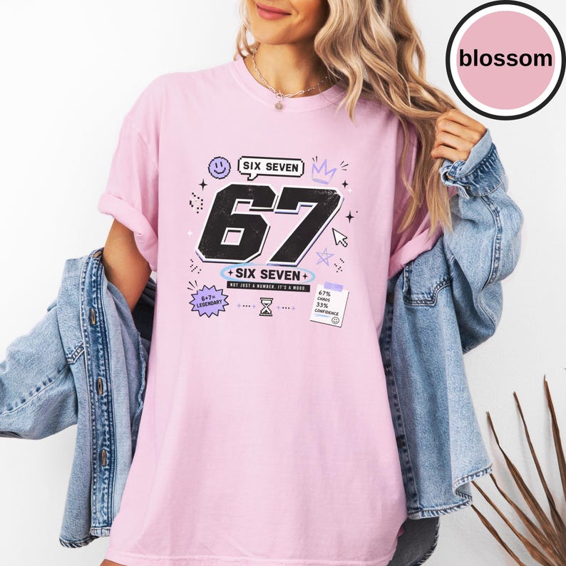 Pu&ograve; includere: Una t-shirt a maniche corte rosa chiaro con una grande grafica nera del numero 67. Il design include il testo SIX SEVEN, NOT JUST A NUMBER. IT'S A MOOD, 6+7= LEGENDARY e 67% CHAOS 33% CONFIDENCE, insieme a piccole icone come una faccina sorridente.