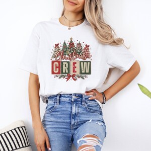 Op de afbeelding: Een wit t-shirt met korte mouwen en een grafische print van verschillende gestileerde kerstbomen met rode strikken. Onder de bomen staat de tekst Christmas CREW in rode en groene blokletters, geaccentueerd met een rode strik en dennentakken.