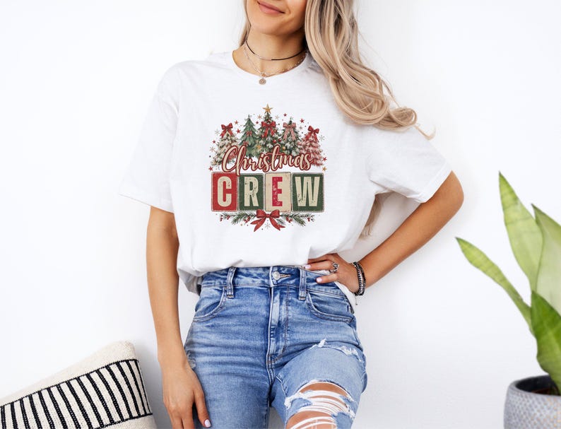 Op de afbeelding: Een wit t-shirt met korte mouwen en een grafische print van verschillende gestileerde kerstbomen met rode strikken. Onder de bomen staat de tekst Christmas CREW in rode en groene blokletters, geaccentueerd met een rode strik en dennentakken.