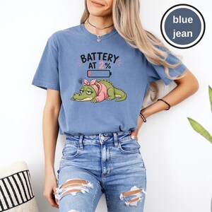 May include: A blue jean coloured t-shirt featuring a graphic of a tired green crocodile wearing a pink headscarf and shirt. Above the crocodile, the text reads BATTERY AT 2% with a low battery icon. The design includes small pink accent marks.