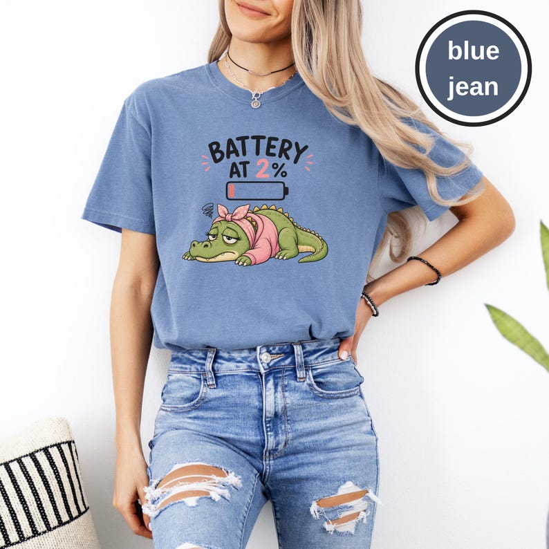 May include: A blue jean coloured t-shirt featuring a graphic of a tired green crocodile wearing a pink headscarf and shirt. Above the crocodile, the text reads BATTERY AT 2% with a low battery icon. The design includes small pink accent marks.