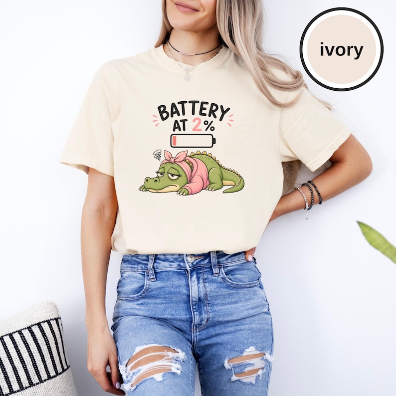 May include: An ivory short-sleeve t-shirt featuring a graphic of a tired green alligator wearing a pink headband. Above the alligator is a battery icon and the text "BATTERY AT 2%" in black lettering. The shirt has a crew neckline and a relaxed fit.