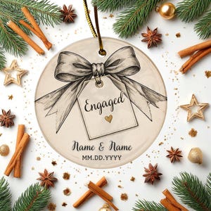 May include: A round ceramic ornament featuring a sketch-style illustration of a large bow tied to a gift tag. The tag displays the text Engaged with a small gold heart, followed by Name & Name and MM.DD.YYYY in black script lettering.