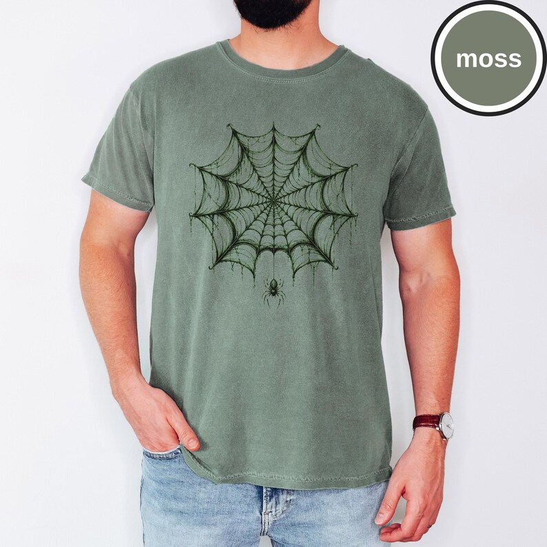 May include: A moss green short-sleeve t-shirt featuring a dark green graphic print of a detailed spider web centered on the chest. A small spider hangs from the bottom of the web. The fabric has a slightly faded, vintage-inspired texture.