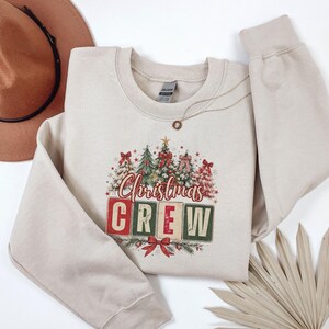 Op de afbeelding: Een beige sweater met ronde hals en een grafische print van verschillende kerstbomen versierd met rode strikken en sterren. Onder de bomen staat de tekst "Christmas CREW" in rode en groene blokletters met een rode strik aan de onderkant.