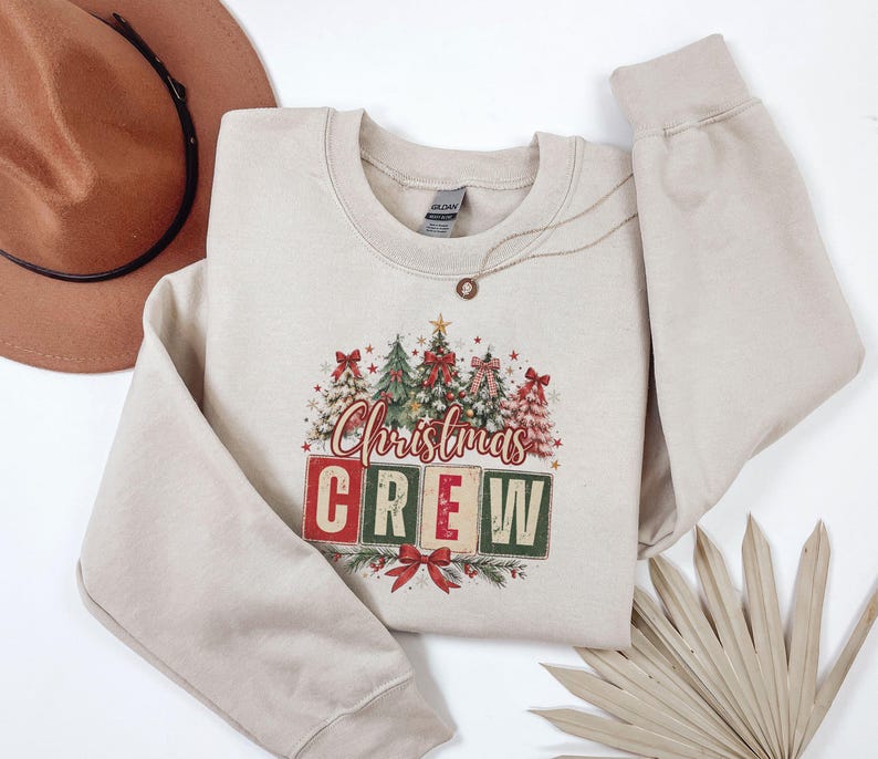 Op de afbeelding: Een beige sweater met ronde hals en een grafische print van verschillende kerstbomen versierd met rode strikken en sterren. Onder de bomen staat de tekst "Christmas CREW" in rode en groene blokletters met een rode strik aan de onderkant.