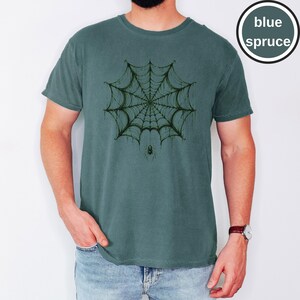 May include: A blue spruce colored short-sleeve t-shirt featuring a dark green graphic print of a spider web with a small spider hanging from the center. The text "blue spruce" appears in a circular badge in the upper right corner of the image.