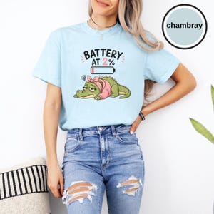 May include: A light blue t-shirt featuring a graphic of a tired green crocodile wearing a pink headband. Above the crocodile, the text reads BATTERY AT 2% with a low battery icon. The shirt is a chambray colour, showcasing a casual short-sleeve design.