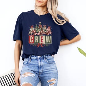 Op de afbeelding: Een marineblauw t-shirt met korte mouwen en een grafische print van verschillende versierde kerstbomen boven de tekst "Christmas CREW" in rode, witte en groene blokletters, geaccentueerd met rode strikken en dennentakken.