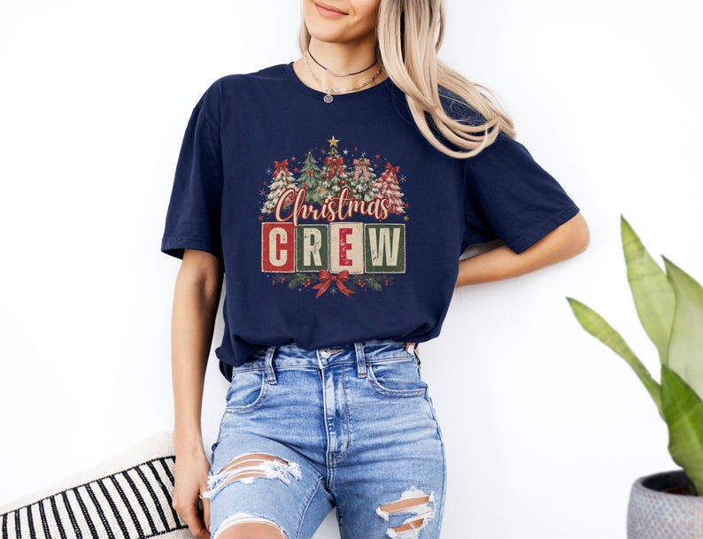 Op de afbeelding: Een marineblauw t-shirt met korte mouwen en een grafische print van verschillende versierde kerstbomen boven de tekst "Christmas CREW" in rode, witte en groene blokletters, geaccentueerd met rode strikken en dennentakken.