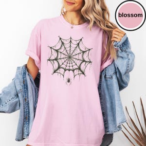May include: A light pink t-shirt featuring a dark green spiderweb graphic centered on the chest with a small spider hanging from the bottom. The shirt has short sleeves and a crew neckline, worn under a blue denim jacket with metal buttons.
