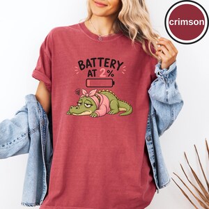 May include: A crimson t-shirt featuring a graphic of a tired green crocodile wearing a pink headscarf and shirt. Above the crocodile is a battery icon and the text BATTERY AT 2%. The shirt is worn with a light blue denim jacket over the top.