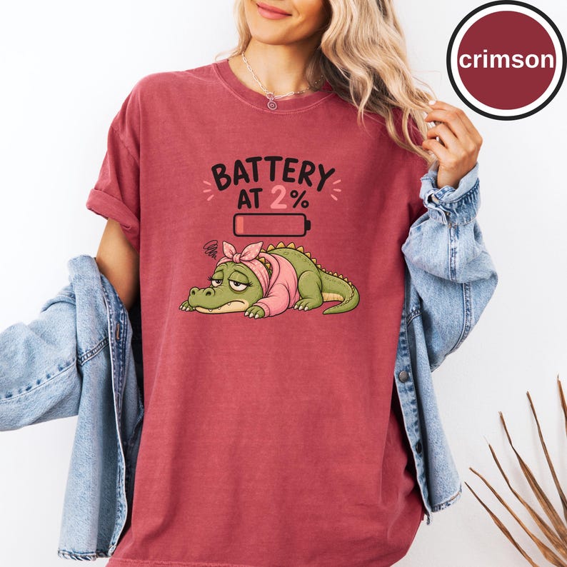 May include: A crimson t-shirt featuring a graphic of a tired green crocodile wearing a pink headscarf and shirt. Above the crocodile is a battery icon and the text BATTERY AT 2%. The shirt is worn with a light blue denim jacket over the top.