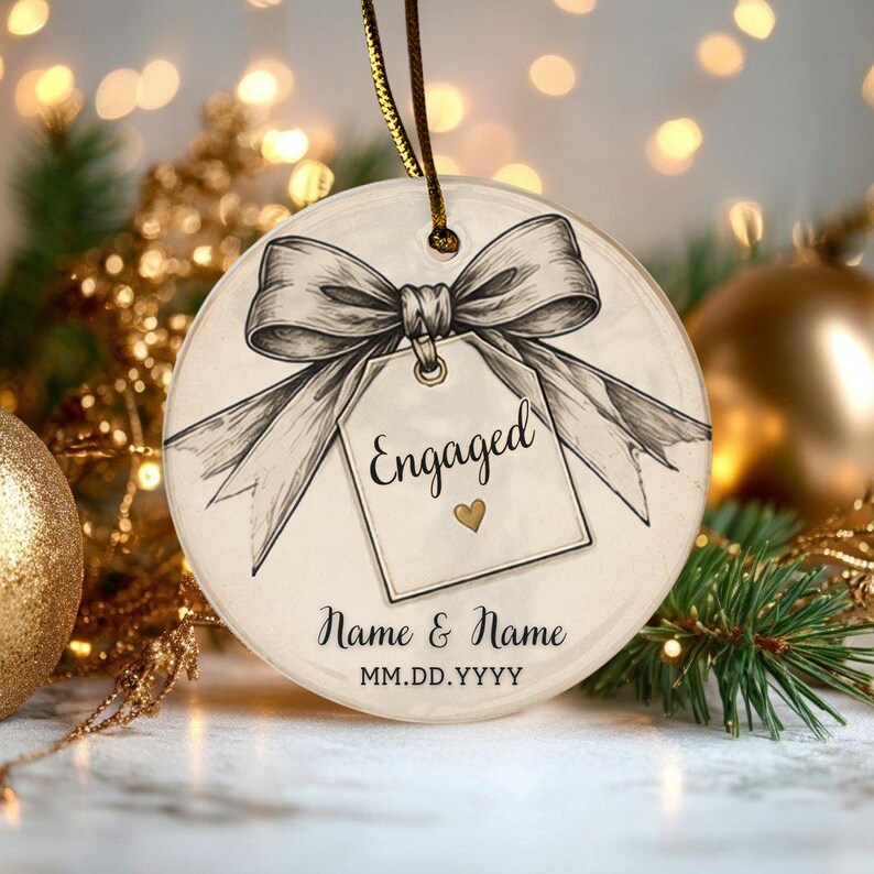May include: A round, clear ornament featuring a black and white sketch of a bow tied to a gift tag. The tag displays the text Engaged with a small gold heart. Below, the text Name & Name and MM.DD.YYYY is printed. A gold string is attached.