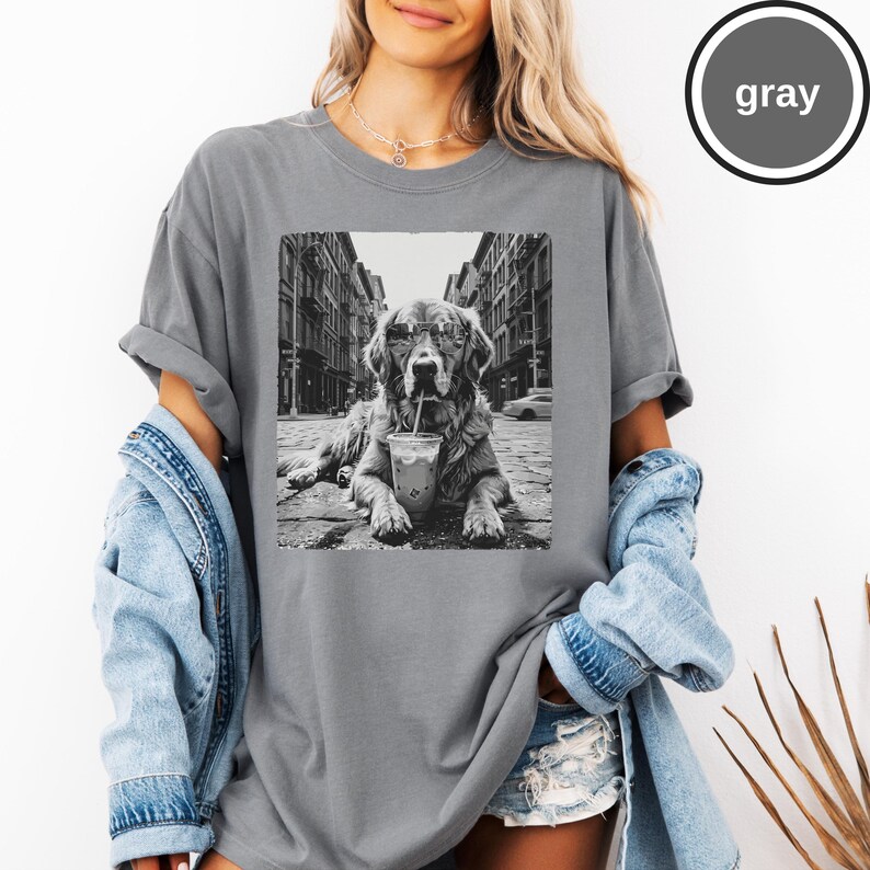 Peut inclure: Un t-shirt gris &agrave; col rond avec une impression graphique en noir et blanc repr&eacute;sentant un chien portant des lunettes de soleil et buvant dans un gobelet en plastique avec une paille, sur fond de rue urbaine. Le t-shirt est port&eacute; sous une veste en jean.