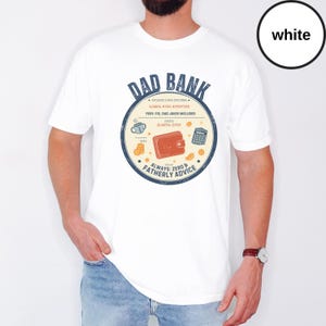 May include: White t-shirt with a circular graphic that reads "DAD BANK" in blue. The graphic includes a wallet, calculator, coffee cup, and coins. The text "ALWAYS: ZERO & FATHERLY ADVICE" is also included.