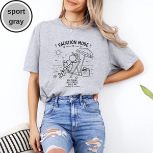 May include: Sport gray t-shirt with a black graphic of a person relaxing on a beach chair under an umbrella, with the text "VACATION MODE" and "NO PLANS. NO STRESS. JUST ME." printed on the front. The shirt is paired with blue jeans.