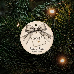 May include: A round white ornament featuring a black and white illustration of a bow tied to a gift tag. The tag displays the text Engaged with a small gold heart. Below the tag, the text Name & Name and MM.DD.YYYY is printed in black ink.