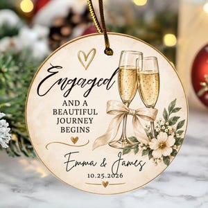 K&ouml;nnte beinhalten: Ein runder Keramikschmuck mit zwei Champagnergl&auml;sern, die mit einem beigen Band verbunden sind, Blumenakzenten und dem Text Engaged AND A BEAUTIFUL JOURNEY BEGINS, Emma & James, und 25.10.2026. Das Design enth&auml;lt goldene Herzen und eine goldene Schnur.