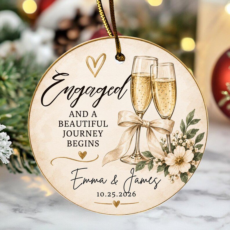 K&ouml;nnte beinhalten: Ein runder Keramikschmuck mit zwei Champagnergl&auml;sern, die mit einem beigen Band verbunden sind, Blumenakzenten und dem Text Engaged AND A BEAUTIFUL JOURNEY BEGINS, Emma & James, und 25.10.2026. Das Design enth&auml;lt goldene Herzen und eine goldene Schnur.
