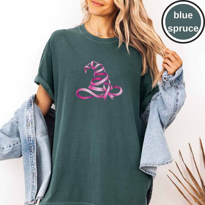 May include: A dark green short-sleeved t-shirt featuring a graphic of a witch hat constructed from a pink ribbon. The shirt has a crew neckline and a relaxed fit. A circular badge in the upper right corner contains the text blue spruce.