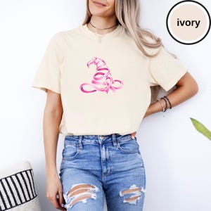 May include: An ivory short-sleeve t-shirt featuring a graphic of a witch hat constructed from a pink awareness ribbon. The shirt has a crew neckline and a relaxed fit. A circular badge in the upper right corner contains the text ivory.