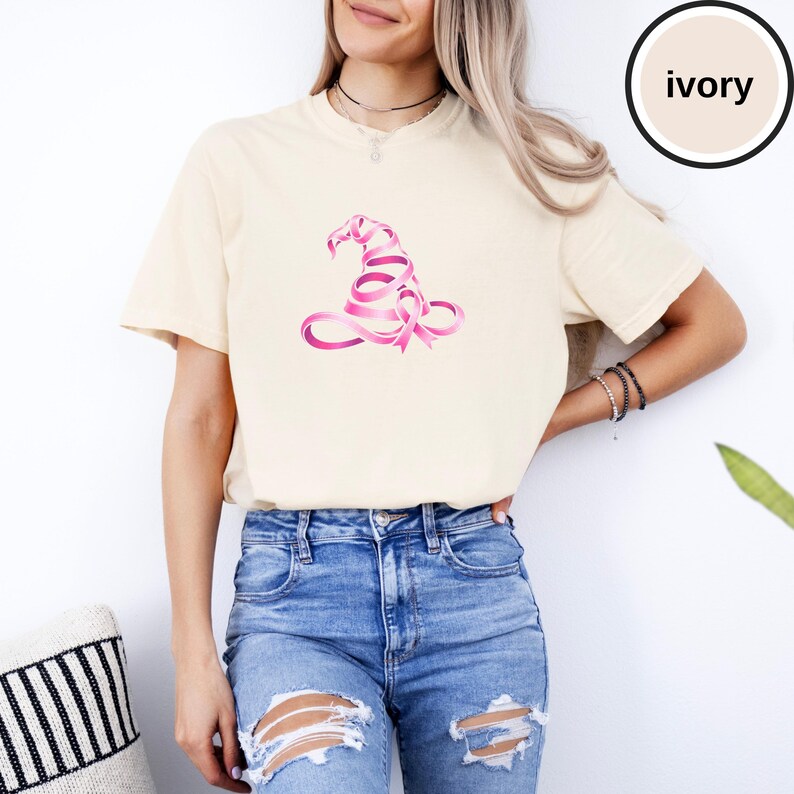 May include: An ivory short-sleeve t-shirt featuring a graphic of a witch hat constructed from a pink awareness ribbon. The shirt has a crew neckline and a relaxed fit. A circular badge in the upper right corner contains the text ivory.