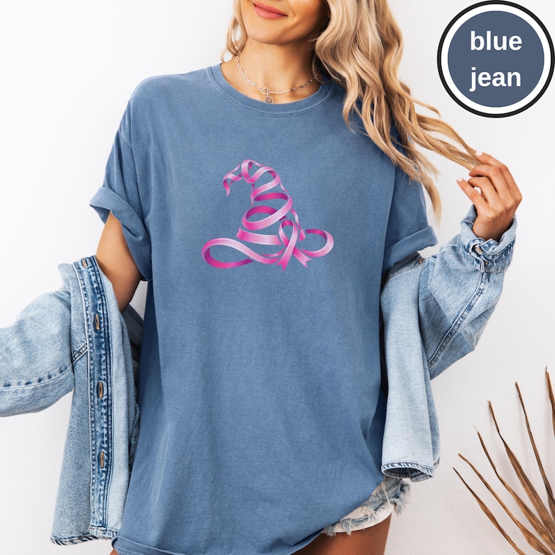 May include: A blue jean colored t-shirt featuring a graphic of a witch hat constructed from a pink ribbon. The shirt has a crew neckline and short sleeves. A circular badge in the upper right corner contains the text blue jean.