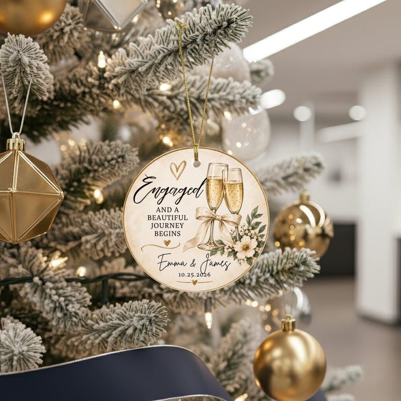 K&ouml;nnte beinhalten: Ein runder, cremefarbener Keramikschmuck mit einer goldenen Schnur. Das Design zeigt zwei Champagnergl&auml;ser, eine Schleife, florale Akzente und den Text: Engaged and a beautiful journey begins, Emma & James, 10.25.2026, mit goldenen Herzen.