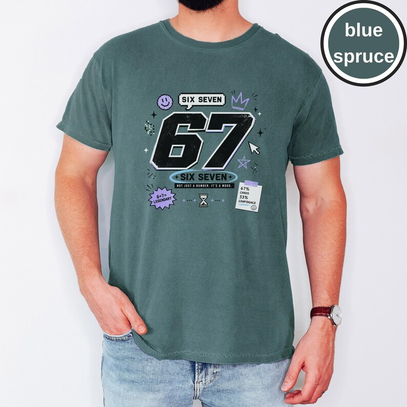 Pu&ograve; includere: Una t-shirt color abete blu con una grande grafica del numero 67 in nero e il testo SIX SEVEN, NOT JUST A NUMBER. IT'S A MOOD. Ulteriori piccole grafiche includono una faccina sorridente, una corona, una stella, una clessidra e una nota con 67% CHAOS 33% CONFIDENCE.