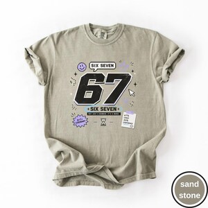 Pu&ograve; includere: Una t-shirt color sabbia con una grafica che presenta il grande numero 67 in nero, il testo SIX SEVEN e frasi aggiuntive tra cui 6+7=LEGENDARY, NOT JUST A NUMBER. IT'S A MOOD, oltre a una nota che riporta 67% CHAOS 33% CONFIDENCE con icone.