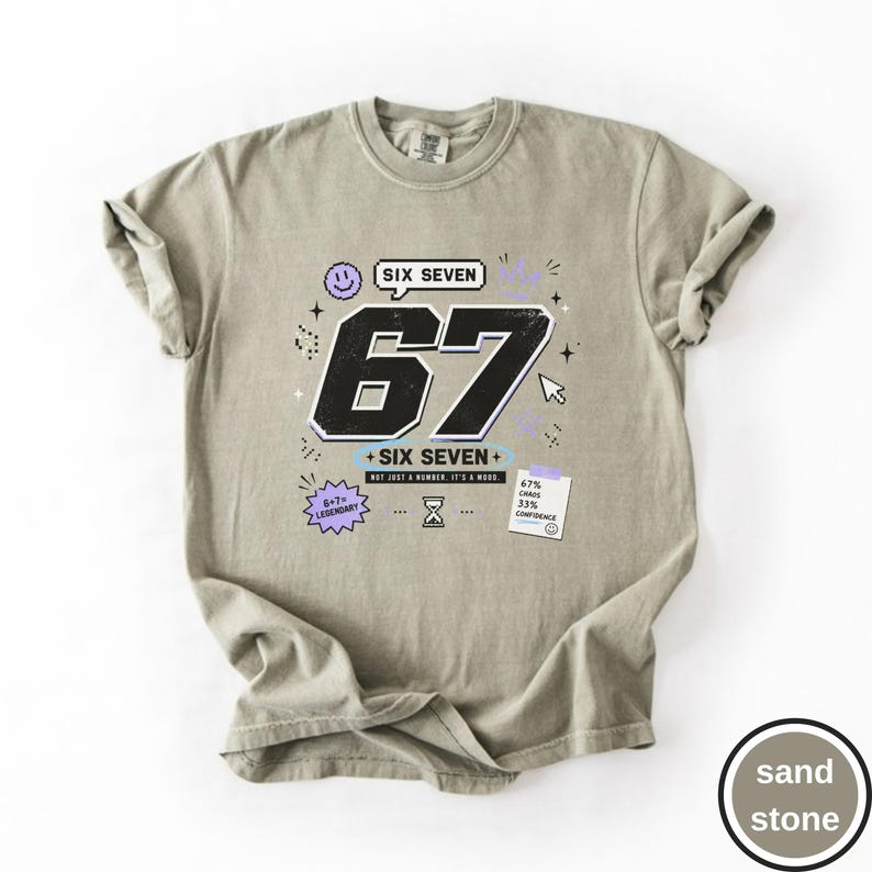 Pu&ograve; includere: Una t-shirt color sabbia con una grafica che presenta il grande numero 67 in nero, il testo SIX SEVEN e frasi aggiuntive tra cui 6+7=LEGENDARY, NOT JUST A NUMBER. IT'S A MOOD, oltre a una nota che riporta 67% CHAOS 33% CONFIDENCE con icone.