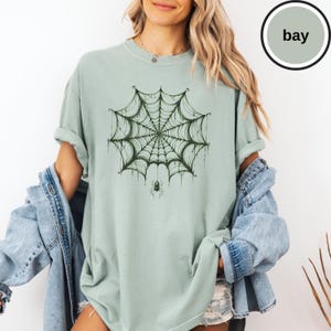 May include: A sage green short-sleeve t-shirt featuring a dark green graphic of a detailed spider web with a small spider hanging from the center. The shirt has a crew neckline and a relaxed fit, layered under a light blue denim jacket.