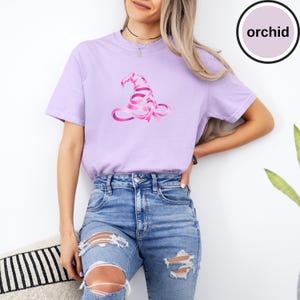 May include: An orchid-colored short-sleeve t-shirt featuring a graphic of a witch hat constructed from a pink awareness ribbon. The shirt has a crew neckline and is paired with blue denim jeans that have distressed rips at the knees.
