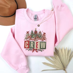 Op de afbeelding: Een lichtroze sweater met ronde hals voorzien van een grafische print van diverse versierde kerstbomen boven de tekst "Christmas CREW" in blokletters, geaccentueerd met rode strikken, sterren en dennentakken, naast een bruine hoed en een gedroogd palmblad.