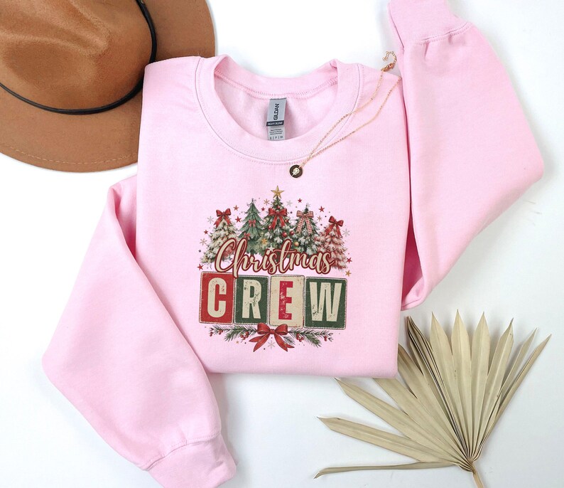 Op de afbeelding: Een lichtroze sweater met ronde hals voorzien van een grafische print van diverse versierde kerstbomen boven de tekst "Christmas CREW" in blokletters, geaccentueerd met rode strikken, sterren en dennentakken, naast een bruine hoed en een gedroogd palmblad.