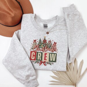 Op de afbeelding: Een lichtgrijs gem&ecirc;leerd sweatshirt met ronde hals voorzien van een afbeelding van diverse versierde kerstbomen en de tekst "Christmas CREW" in rode en groene blokletters, geaccentueerd met rode strikken en kleine sterretjes, met een bruine hoed.