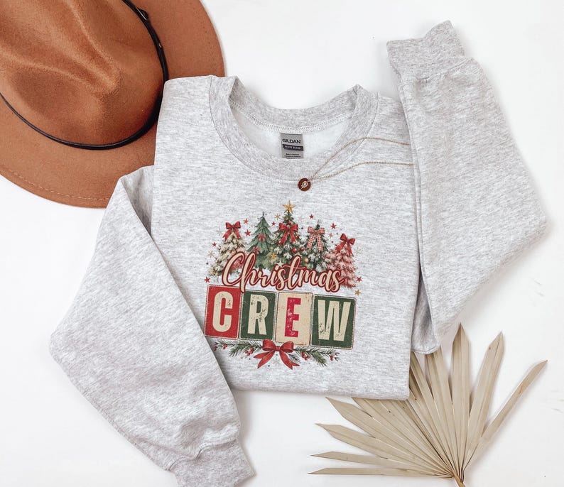 Op de afbeelding: Een lichtgrijs gem&ecirc;leerd sweatshirt met ronde hals voorzien van een afbeelding van diverse versierde kerstbomen en de tekst "Christmas CREW" in rode en groene blokletters, geaccentueerd met rode strikken en kleine sterretjes, met een bruine hoed.